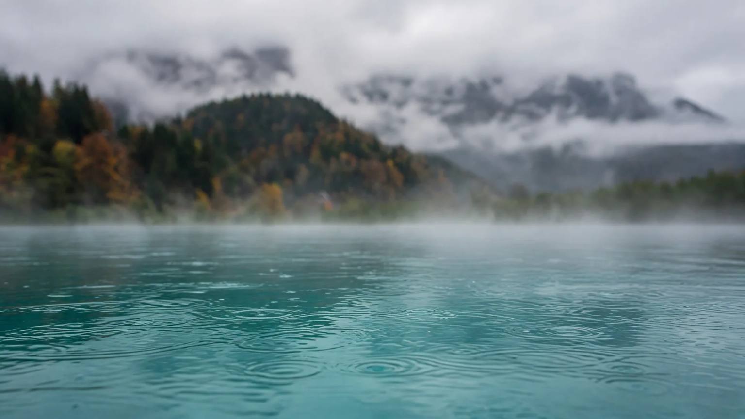 Dampfendes, türkises Außenbecken an einem Regentag im Allgäu, dahinter herbstlich bewaldete Hügel und Berge in tiefen Wolken