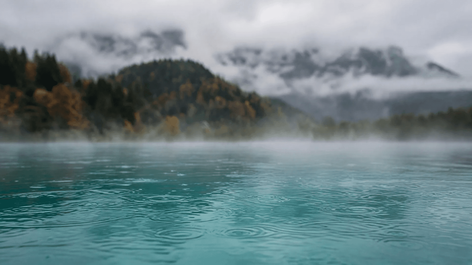 Dampfendes, türkises Außenbecken an einem Regentag im Allgäu, dahinter herbstlich bewaldete Hügel und Berge in tiefen Wolken