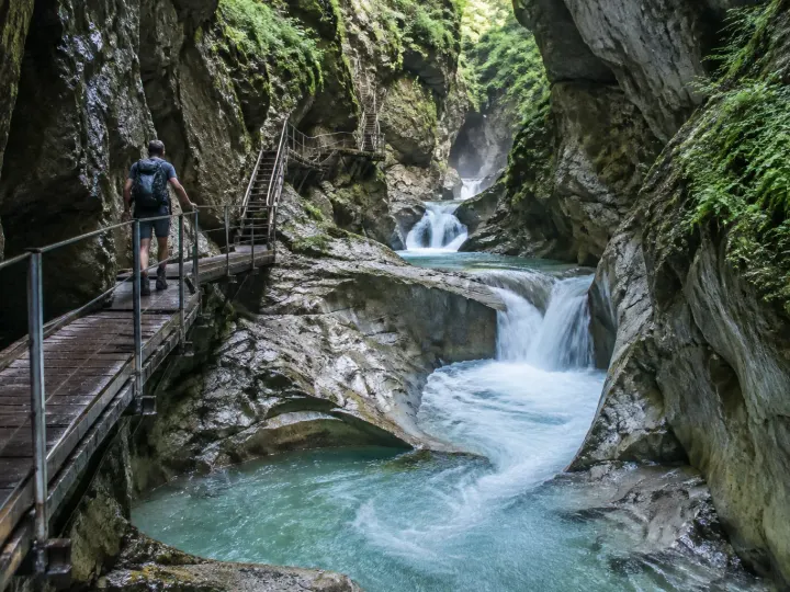 Wanderer auf dem Holzsteg der Starzlachklamm bei Sonthofen, darunter stürzt türkisfarbenes Wasser zwischen moosigen Felswänden