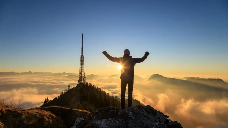Person mit erhobenen Armen auf dem Grünten-Gipfel bei Sonnenuntergang über dem Allgäu
