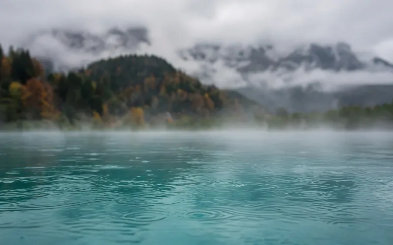 Dampfendes, türkises Außenbecken an einem Regentag im Allgäu, dahinter herbstlich bewaldete Hügel und Berge in tiefen Wolken