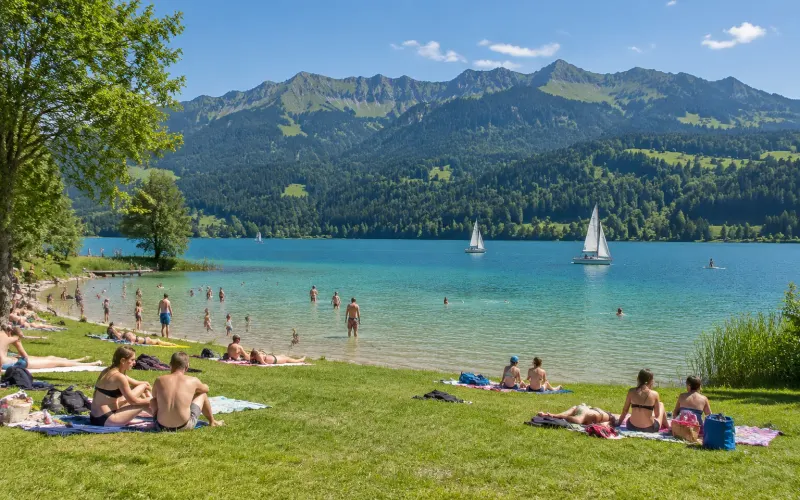 Badewiese am Großen Alpsee bei Immenstadt: Menschen liegen in der Sonne und baden im türkisen Wasser, Segelboote vor den bewaldeten Bergen