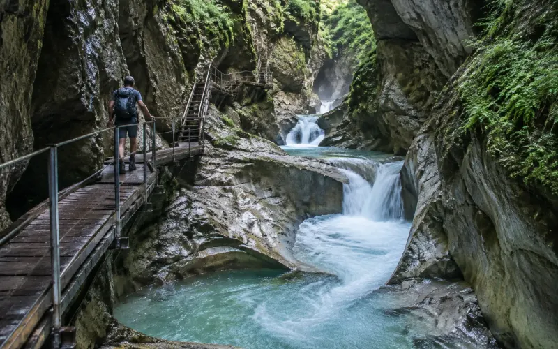Wanderer auf dem Holzsteg der Starzlachklamm bei Sonthofen, darunter stürzt türkisfarbenes Wasser zwischen moosigen Felswänden