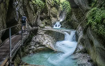 Wanderer auf dem Holzsteg der Starzlachklamm bei Sonthofen, darunter stürzt türkisfarbenes Wasser zwischen moosigen Felswänden